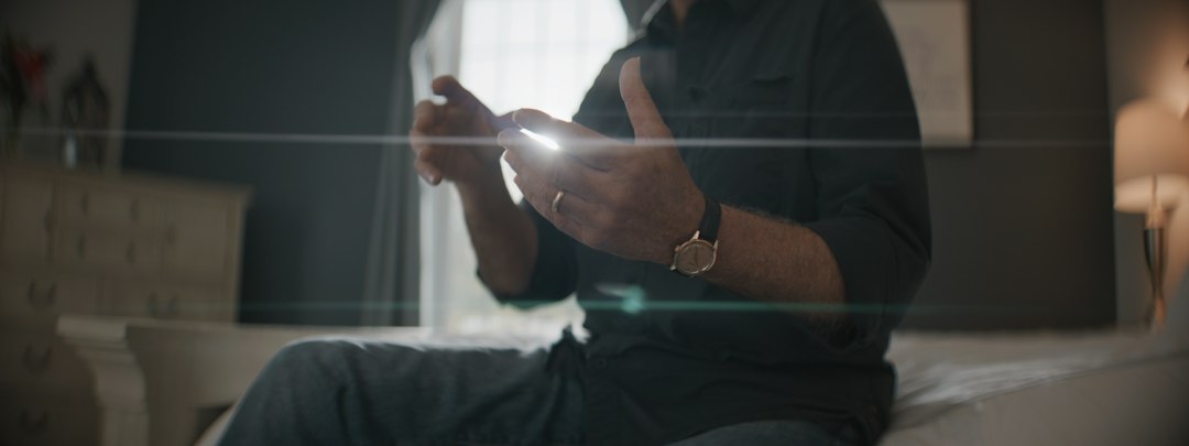 寝る前スマホがやめられない人 — ベッドに腰かけて、光る画面のスマートフォンを操作する手元